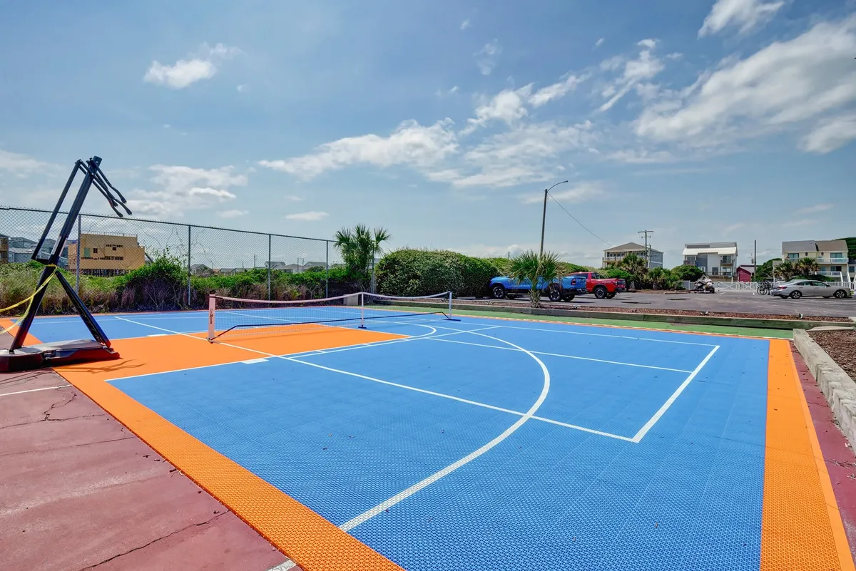 The scene features a vibrant outdoor basketball and tennis court, complete with a basketball hoop, tennis net, and a colorful court surface under sunny skies. The area is designed for friendly matches and group activities, offering plenty of space for social gatherings and exercise.