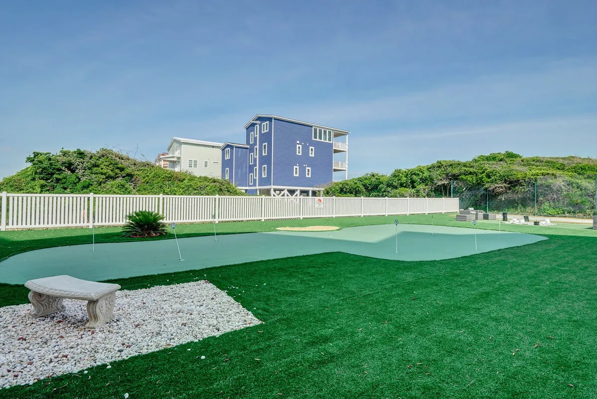 An exterior scene featuring a lush artificial putting green, a stone bench, mature landscaping, and a glimpse of a blue house. The ambiance is tranquil and inviting, suggesting leisurely outdoor activities as a couple, such as relaxing on the bench or enjoying gentle strolls at sunset.