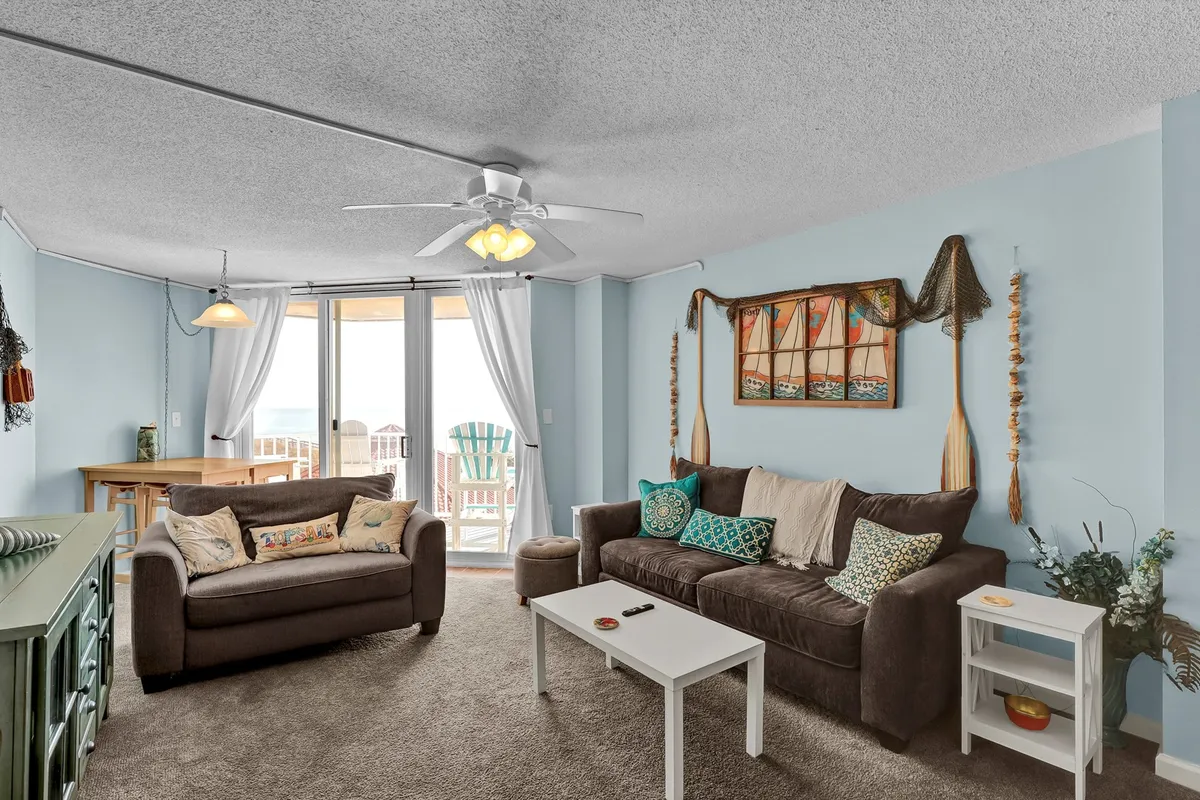 A cozy living room with a comfortable sofa and a dining table sits in front of broad balcony doors, revealing ocean views beyond. Tasteful artwork and warm wood tones add to the inviting, casual ambiance, suggesting both relaxation and shared meals with friends and family.