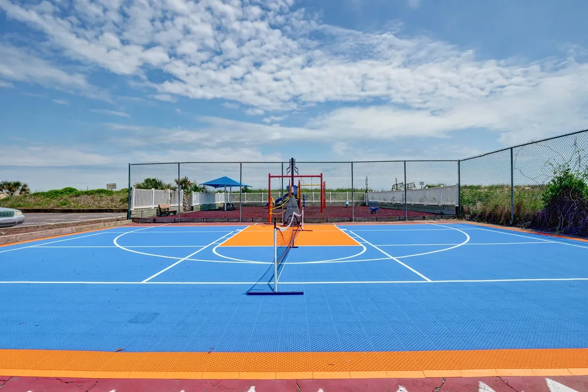 A lively outdoor recreation area with colorful basketball and tennis courts, playground equipment, and surrounding greenery. Children are seen playing, while families and friends gather for active fun under the sun.