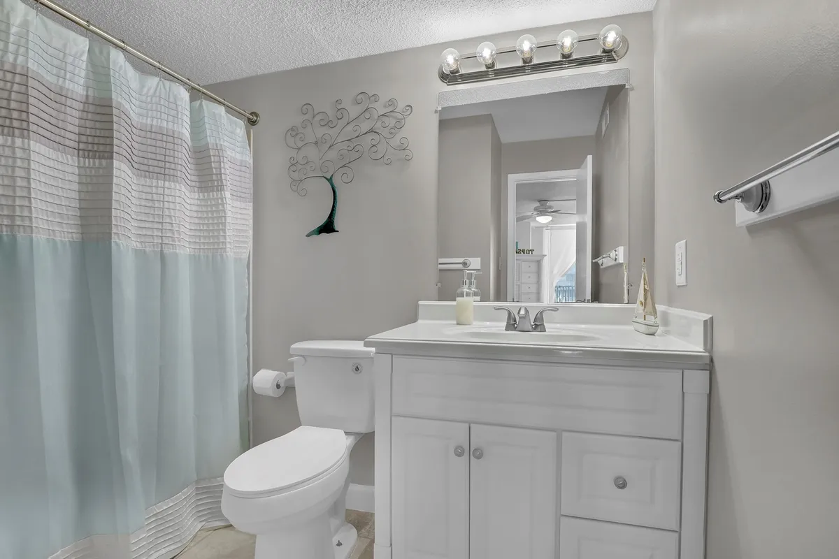 A bright, inviting bathroom featuring a white vanity with a large mirror and stylish light fixtures, decorative wall art, and a neatly arranged shower area. The feel is fresh, clean, and modern.