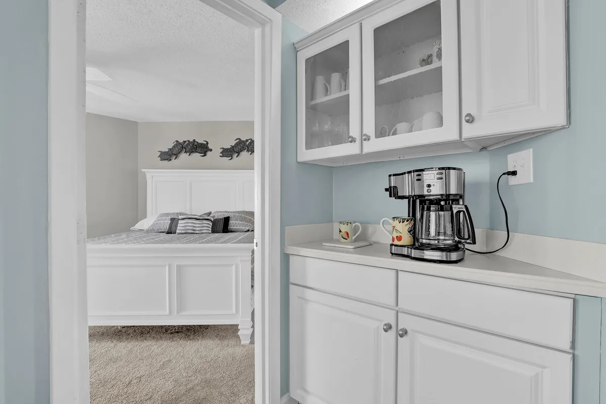 A compact coffee nook featuring a modern coffee maker set against white cabinetry. Through the adjacent doorway, a glimpse of the bedroom is visible, adding a sense of intimacy and warmth. Morning light filters in, enhancing the cozy, inviting feel ideal for starting the day.