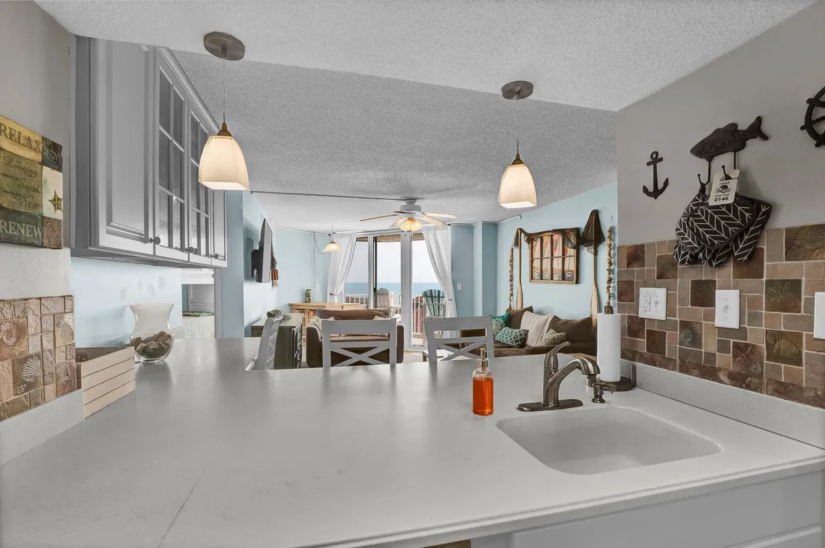 An inviting open-plan space with a kitchen island, adjacent dining area, decorative wall features, and large windows that offer a glimpse of the ocean view. The setup is perfect for group gatherings, casual breakfasts, or holiday celebrations.