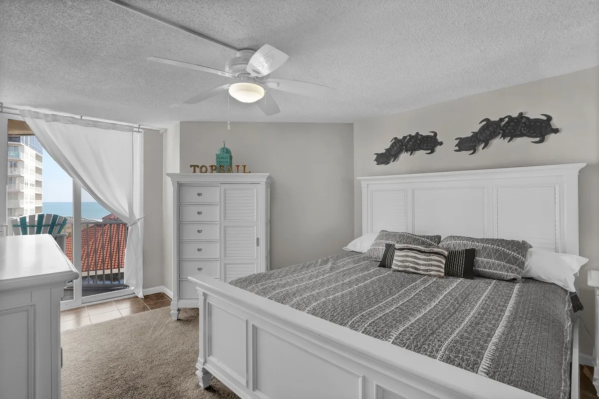 A serene master bedroom featuring a plush bed with textured bedding, a white dresser, and a window that offers a glimpse of the ocean outside. The decor is calm and coastal, creating a restful atmosphere ideal for relaxation.