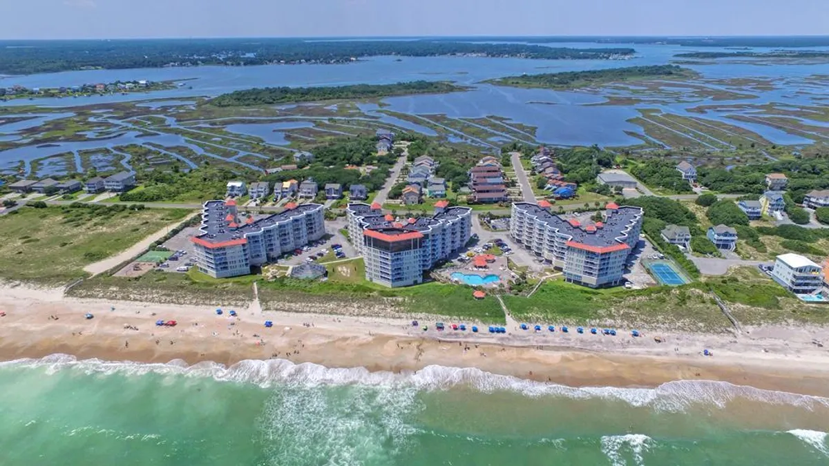 A vibrant exterior shot capturing beachfront resort buildings, the sparkling outdoor swimming pool, sandy shoreline, and lush dune vegetation. Guests are seen swimming in the pool, sunbathing, playing beach volleyball, and strolling along the shore, evoking a lively and relaxed resort atmosphere.