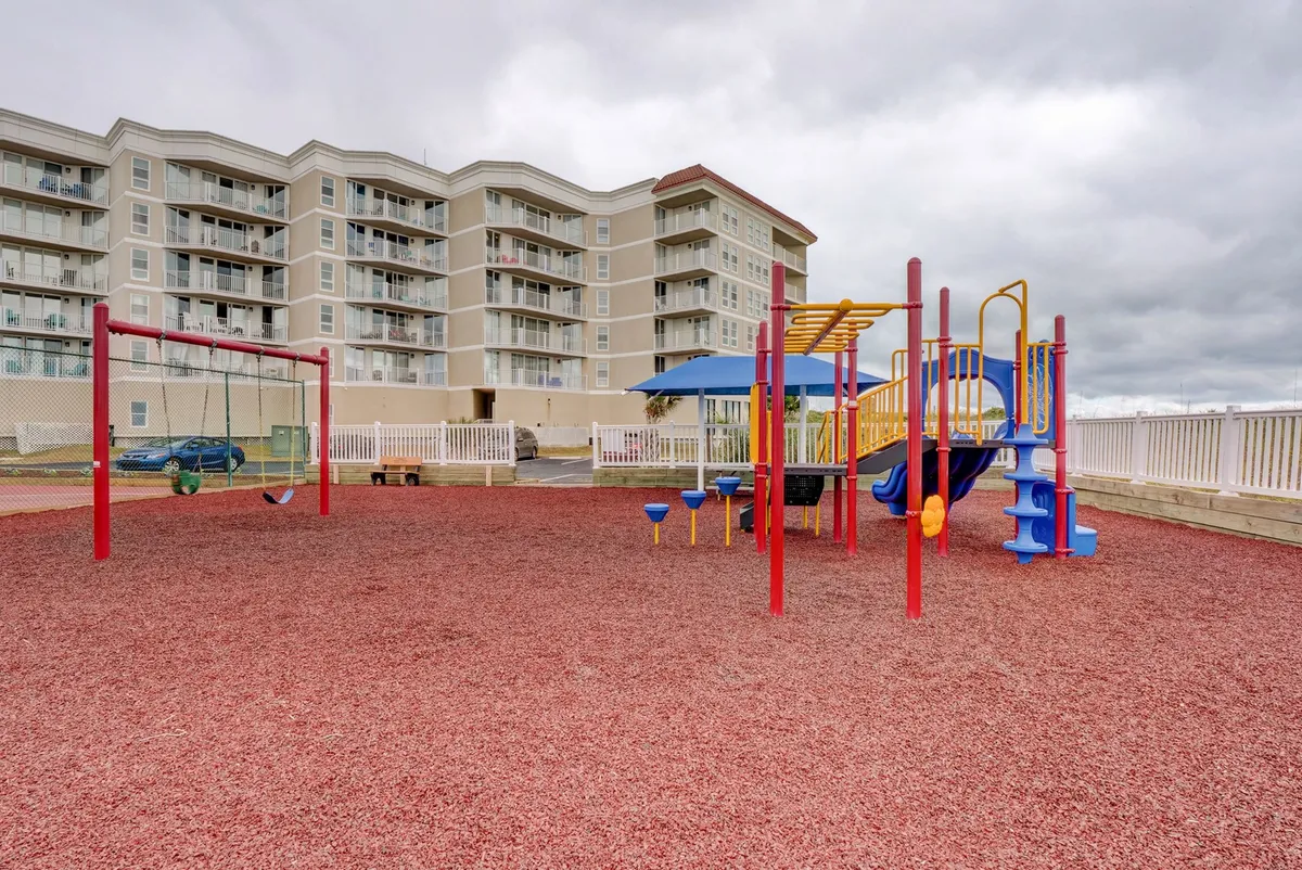 A cheerful playground scene featuring swings, bright red rubber mulch, and a blue slide, with children actively playing and parents socializing nearby. The area exudes vibrancy and safety, representing a key amenity for families with young children.