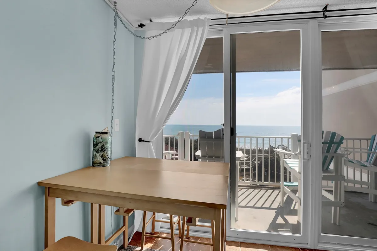A bright, open-concept living and dining area with a wooden table sits beside large sliding doors that offer sweeping views of the ocean. Light curtains billow gently, hinting at a fresh sea breeze. The space feels tranquil and welcoming, ideal for both relaxing and enjoying meals with a view.