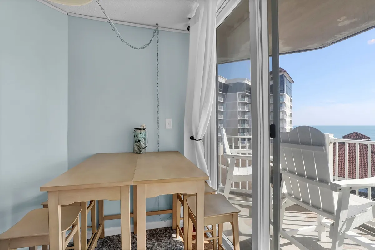 A wooden dining table with chairs positioned beside a large window, framing a stunning ocean view. A decorative vase sits atop the table, setting a relaxed and intimate dining mood.