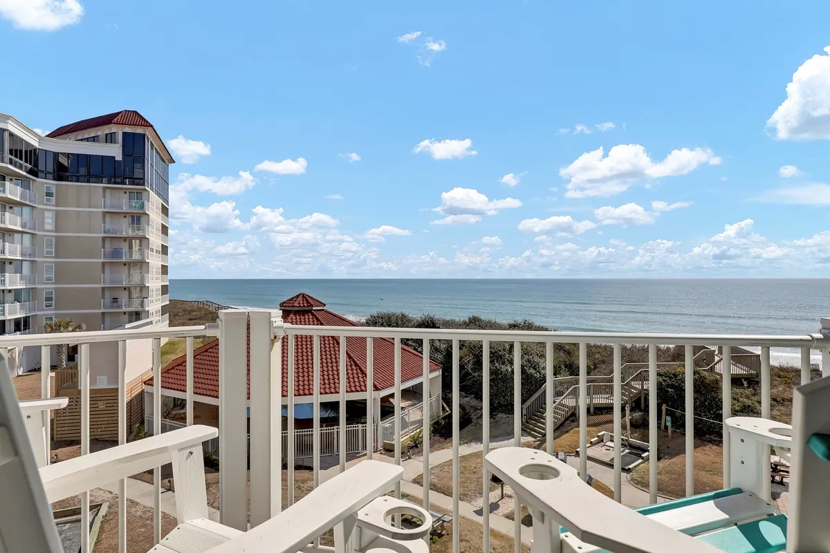 A sweeping view from a balcony, capturing the ocean, nearby buildings, and the balcony railing. The scene is peaceful and inviting, evoking the sense of waking up to a beautiful day by the coast or enjoying the calm at the day's end. The view complements activities like sharing a meal, barbecuing, or simply enjoying the ambiance.