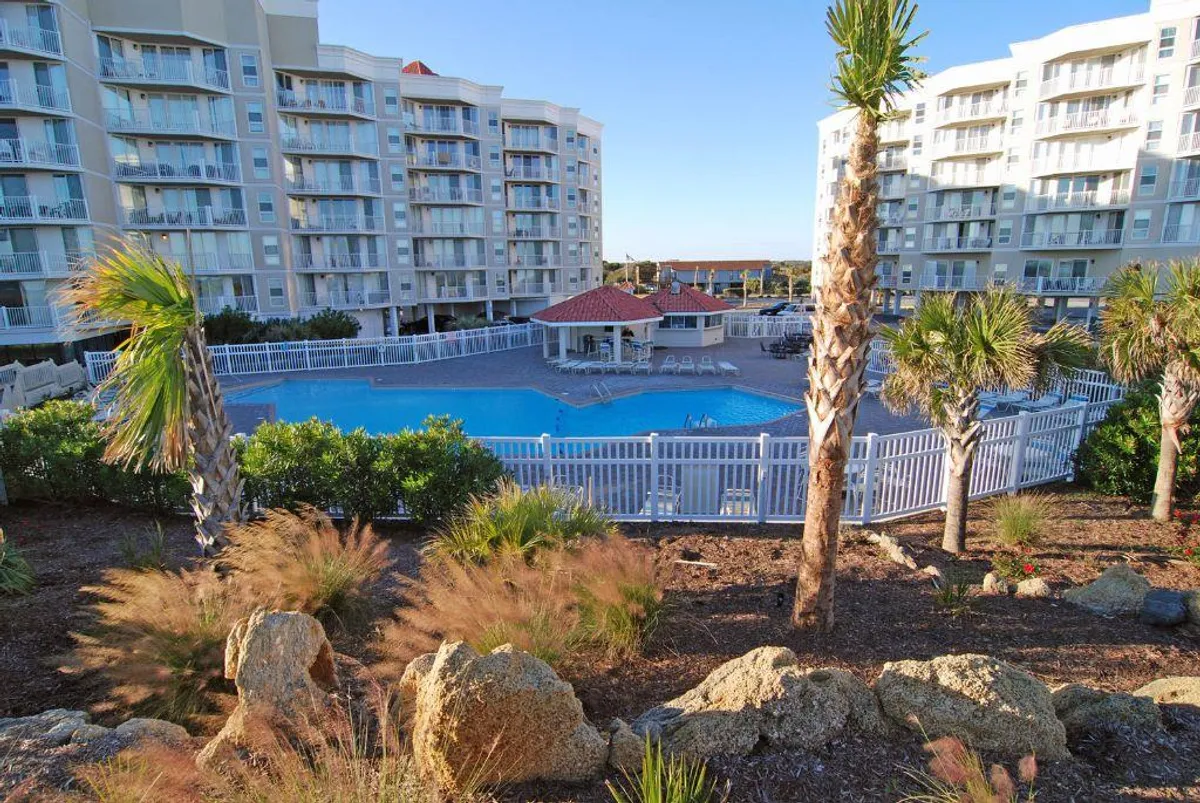 A vibrant outdoor pool scene surrounded by palm trees and modern condominium buildings, with lounge chairs and a pavilion featuring a red roof. The tranquil water and welcoming atmosphere invite guests to relax, swim, or socialize poolside.